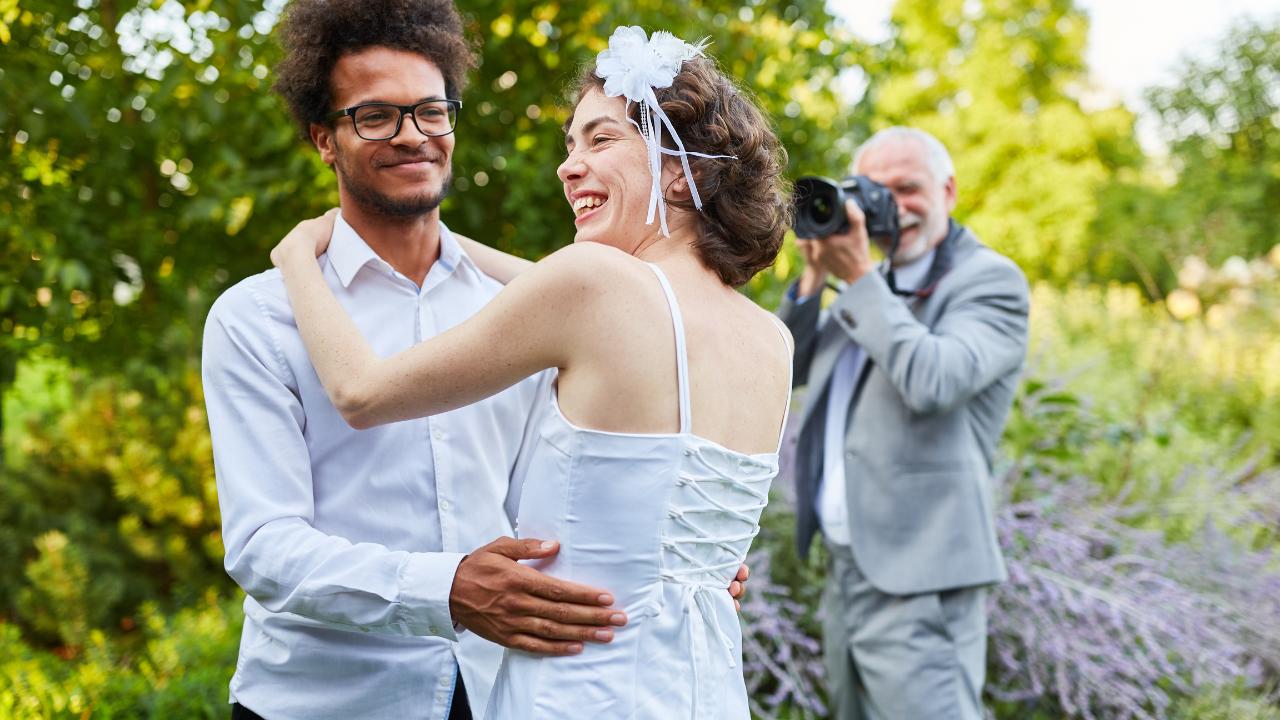 Casal de noivos, composto por homem com óculos de grau preto e camisa branca, e mulher com vestido e flor no cabelo brancos. Os dois abraçados e sorrindo naturalmente. Ao fundo, o fotógrafo, vestido com terno e calça cinzas e blusa branca, registra o momento em ensaio pré-wedding.