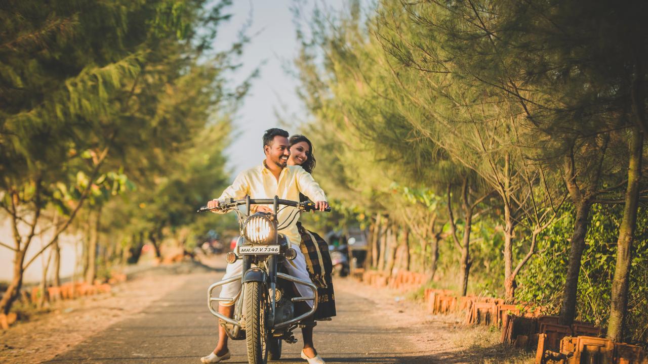 Homem com camisa amarela, calça e sapatos brancos, com mulher sentada atrás dele em motocicleta  parada no meio da estrada, com árvores nas margens. Ambos estão sorrindo.