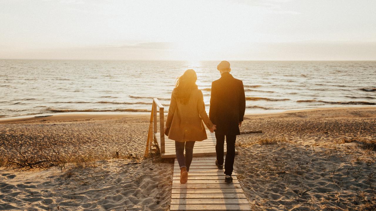 Casal andando de costas em uma ponte na areia da praia com o mar ao fundo.