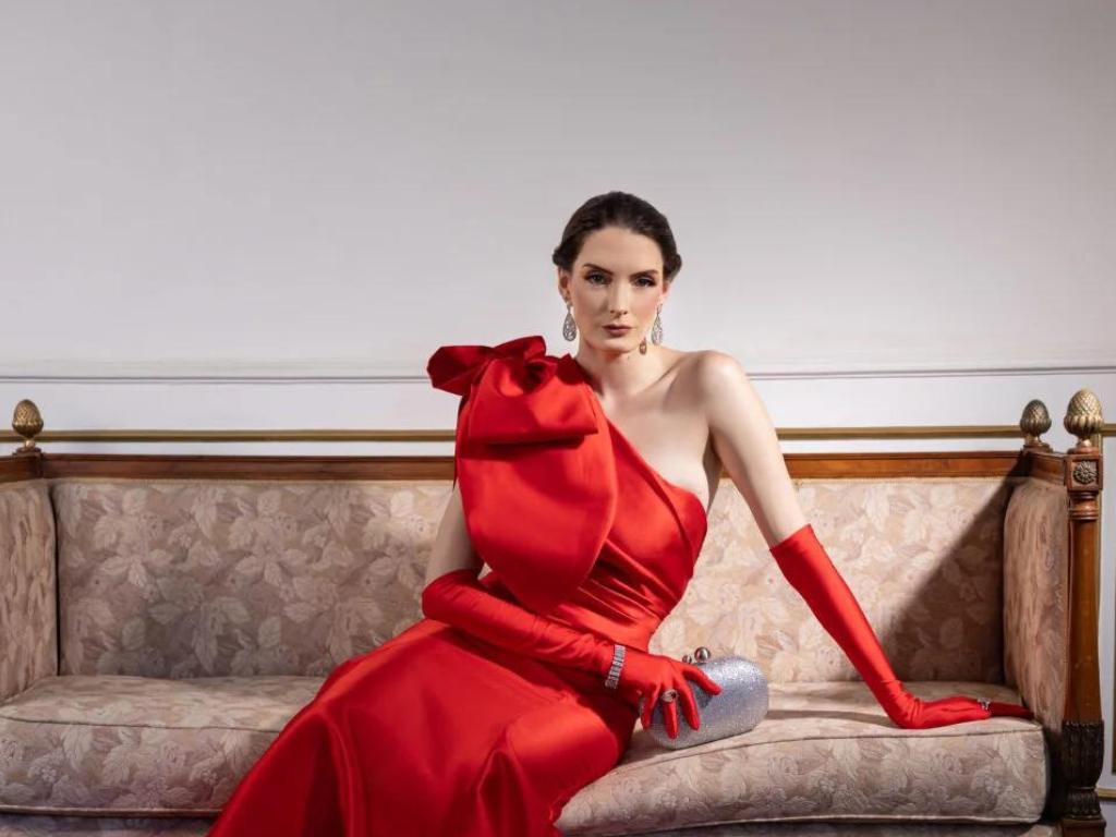 mulher sentada em um sofá com um dos ombros apoiado vestindo vestido vermelho um ombro só com laço grande em um dos lados e luvas vermelhas