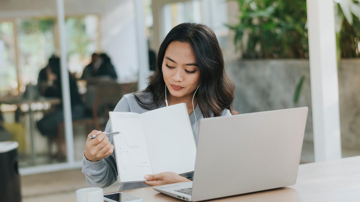 mulher com fones de ouvido em frente ao notebook apontando para um caderno