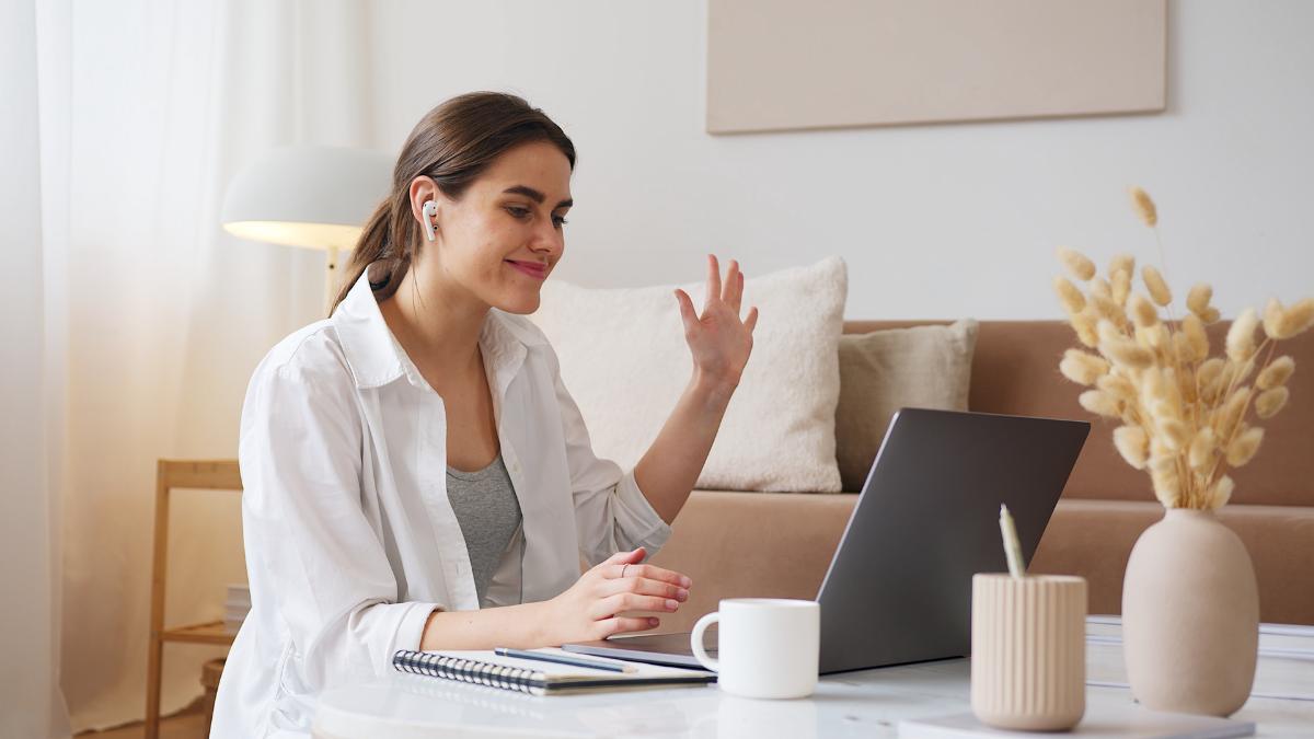 mulher em uma sala com camisa branca e fone acenando para o computador