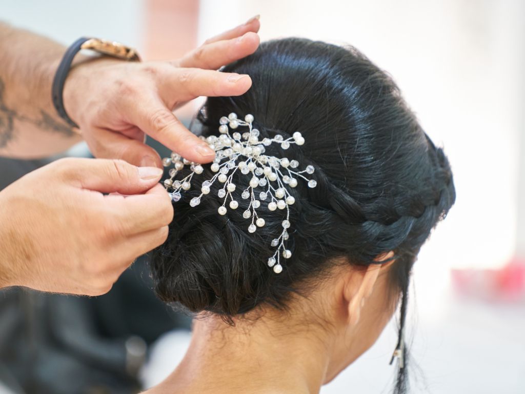 mulher de cabelo preto com um coque com trança frontal e acessório com pérolas pequenas sendo presa por uma pessoa